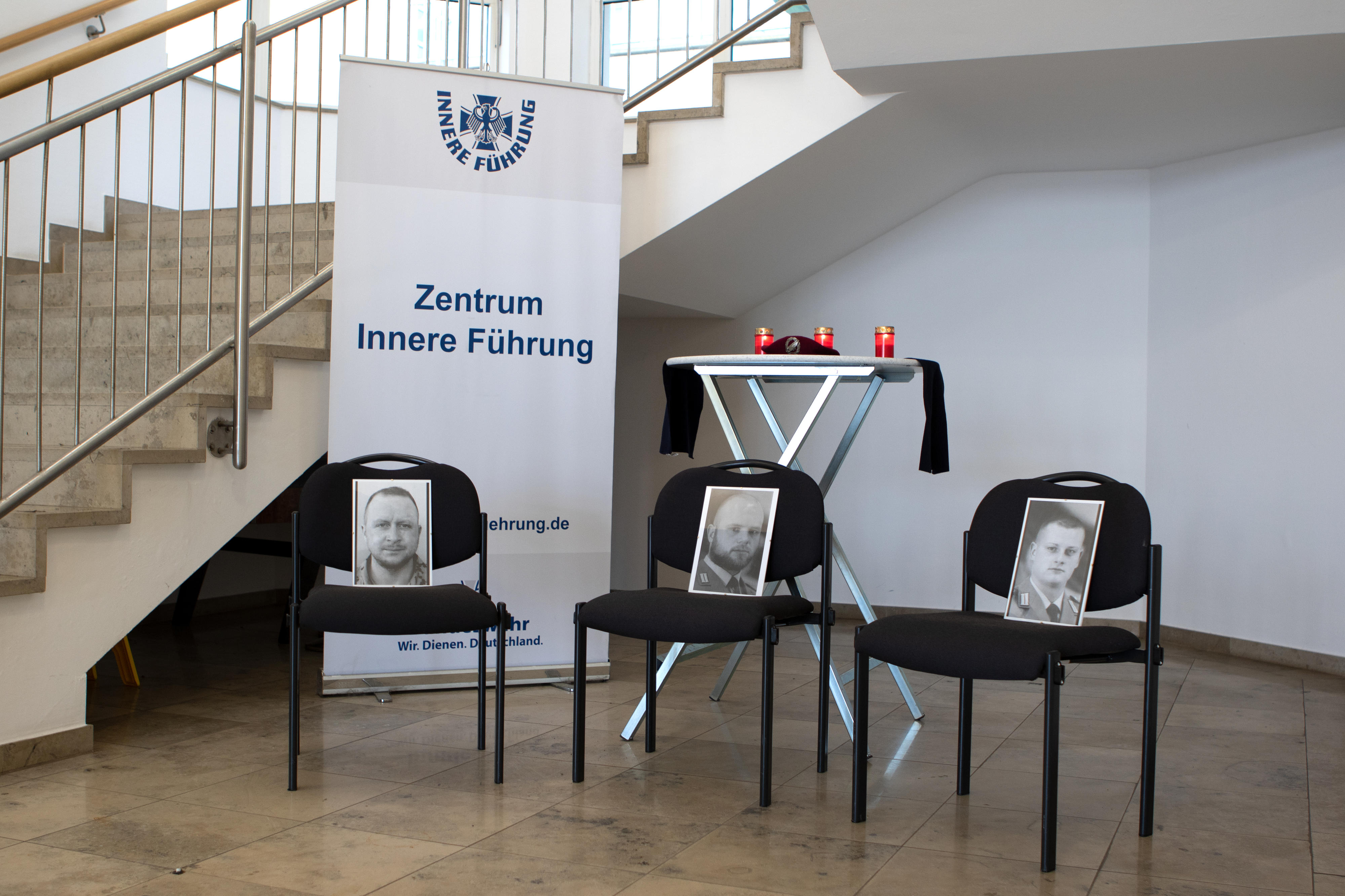 Drei leere Stühle mit Porträtfotos dreier Gefallener der Bundeswehr, vor drei Kerzen auf einem Stehtisch. 