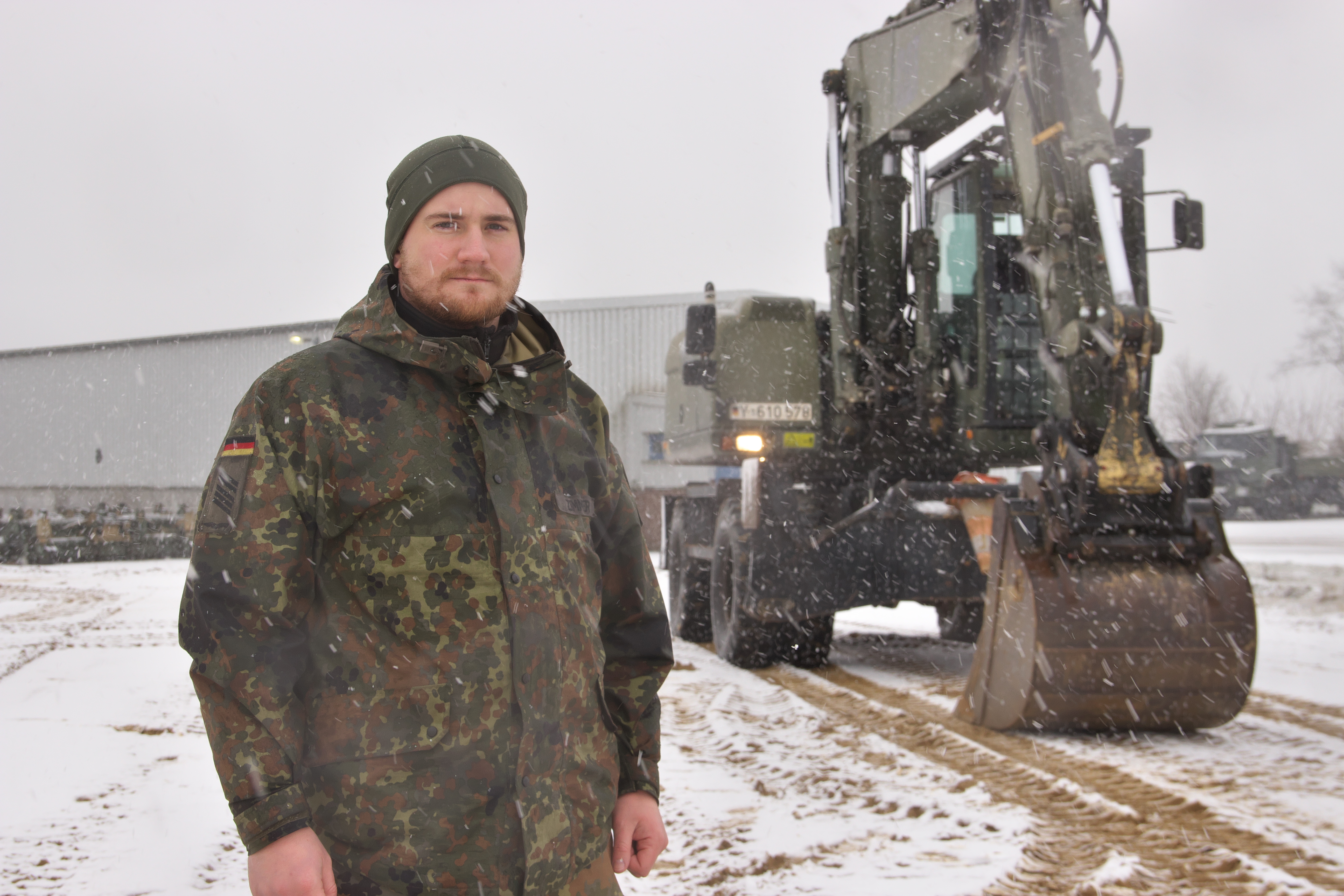 Ein Soldat schaut in die Kamera; rechts hinter ihm steht ein Bagger.