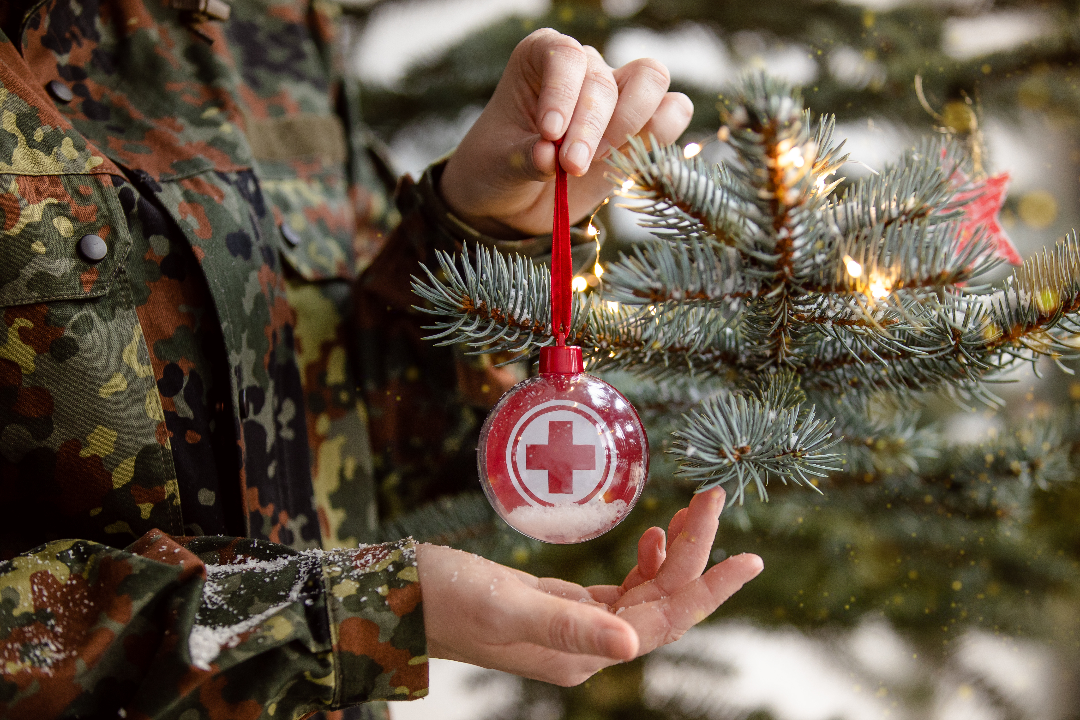 Soldatin hält eine Kugel mit einem rotem Kreuz vor einem Weihnachtsbaum