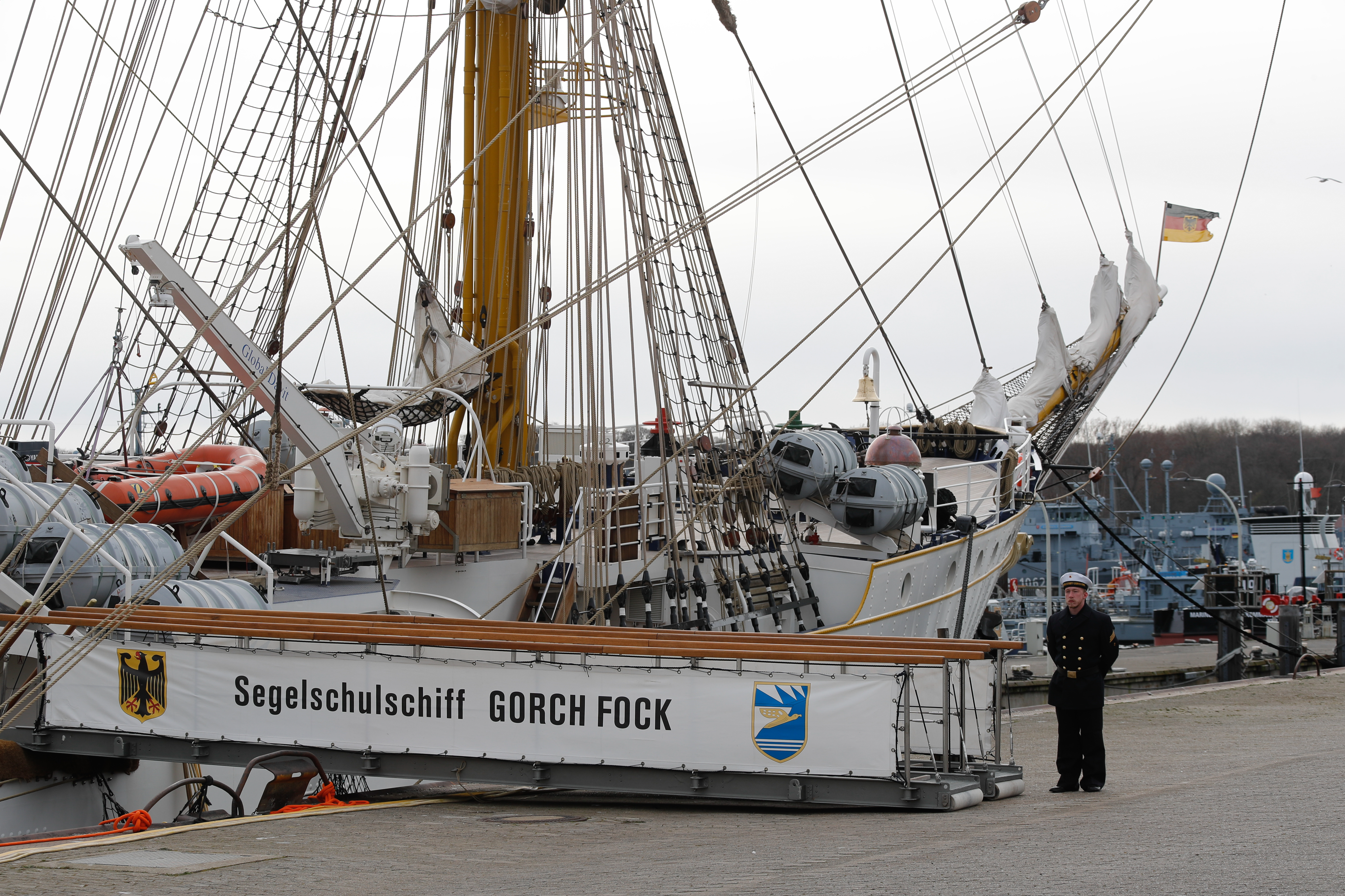 Ein großes weißes Segelschiff liegt im Hafen. Ein Stelling führt von Bord vor der ein Mann in Uniform steht.