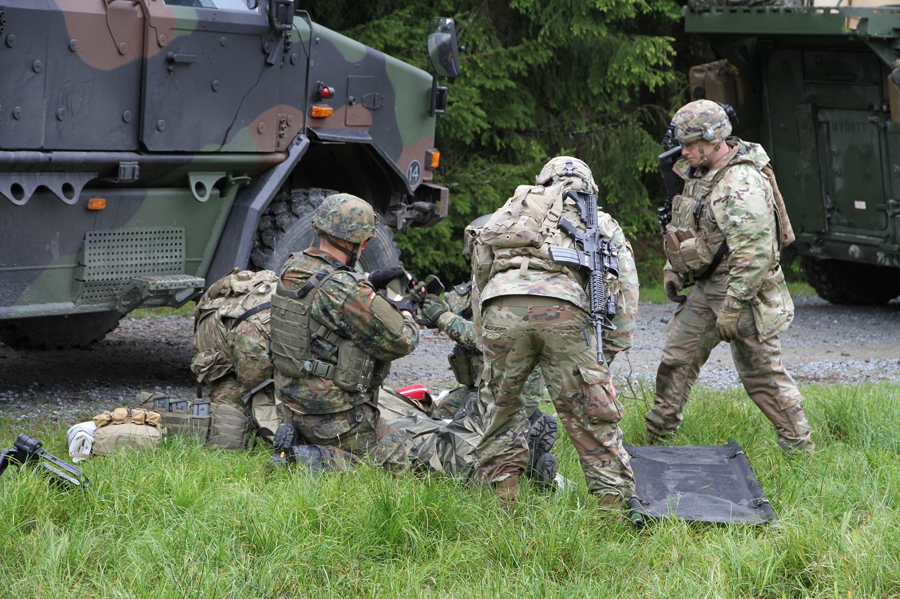 Vor einem Fahrzeug stehen und hocken mehrere Soldaten um einen Verwundeten am Boden herum.