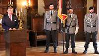 Ein Mann steht in einem historischen Saal an einem Rednerpult, neben ihm Soldaten mit Truppenfahne.