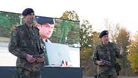 Zwei Soldaten mit Headset stehen auf einer Bühne, hinter ihnen ein Großleinwand. 