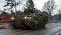 Ein mit Ästen getarnter Schützenpanzer Marder fährt auf einer Landstraße an Häusern vorbei.