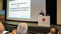Ein Mann hinter einem Rednerpult neben einer großen Leinwand mit einem Willkommensgruß