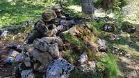 Zwei Soldaten sitzen hinter einem Erd- und Steinwall mit dem Gewehr im Anschlag.