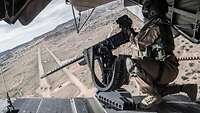Bei geöffneter Heckklappe überfliegt der Hubschrauber eine Freifläche und eine Soldatin kniet hinter der Waffe.