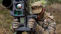 Ein Soldat sitzt in einem Wald und beobachtet durch ein Lenkflugkörpersystem das vor ihm liegende Waldstück.