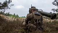 Ein Soldat beobachtet in einem Waldstück durch das Visier des Systems MELLS das vor ihm liegende Gelände.