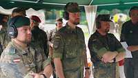 Drei Soldaten mit Gehörschutz stehen unter einem Zeltdach nebeneinander.
