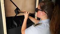 Soldatin im Dienstanzug spielt auf einer Oboe, die durch eine Holzblende durch einer Wand gesteckt ist.