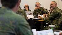 Mehrere Soldaten sitzen um einen Tisch herum, lächeln und haben Kopfhörer auf.