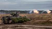 Ein Panzer fährt über einen Gefechtsplatz, im Hintergrund Rauch und Feuer von Munition.