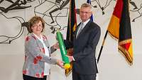 Weibliche Person übergibt überdimensionalen Stift mit grüner Farbe an männliche Person