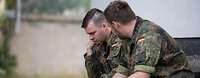 Zwei Soldaten sitzten leicht gekrümmt vor einem Gebäude auf einer Holzbank und unterhalten sich