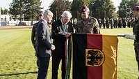 Zwei Zivilisten und ein Soldat stehen an der Truppenfahne, ein Zivilist befestigt ein Fahnenband.