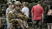 Ein bewaffneter Soldat mit Helm, Mundschutz und Sonnenbrille, hinter ihm zivile Besucher.