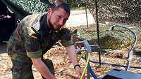 Soldat mit Messbecher an einem Wasserhahn, im Hintergrund ein Zelt und sandiger Untergrund.
