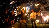 Drei Frauen und ein Mann stehen nebeneinander und halten Feuerwerke an Stangen in den Händen.