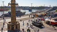 Blick von oben auf ein Volksfest am Hafen, mit Imbissbuden, Fahrgeschäften und einem Schiff am Kai.