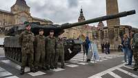 Vier Soldaten stehen nebeneinander vor einem Panzer, der auf einem Stadtplatz steht.