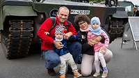 Ein Vater und eine Mutter hocken mit ihren zwei kleinen Kindern vor einem Militärfahrzeug.