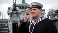 Ein Matrose steht vor einem Marineboot. Er hebt den Arm und hat eine Signalpfeife in der Hand.