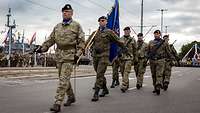 Sechs Soldaten marschieren mit der Korpsfahne über eine breite Straße, die von Menschen gesäumt ist.