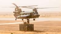 Ein Hubschrauber im Schwebeflug. Unter ihm steht ein Container, auf dem sich Soldaten befinden 