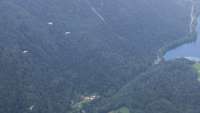 Drei Soldaten am Fallschirm fliegen über einen Talkessel, im Hintergrund ist ein See zu sehen.