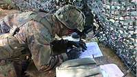 Ein Soldat trägt Daten in ein Formular ein. Er hockt in einem Erdloch, davor ein Tarnnetz gespannt. 