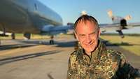 Portraitaufnahme eines Offiziers in Flecktarnuniform; im Hintergrund ein großes, viermotoriges Propellerflugzeug.