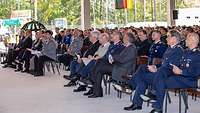 Viele Personen, davon einige in Uniform, auf Stühlen in einer Halle.