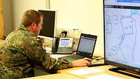 Rechts ein Bildschirm mit Karte. Daneben sitzt ein Soldat und bedient einen Laptop.