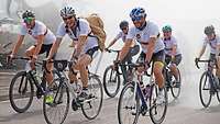 Eine Gruppe Radfahrer fährt durch einem Sprühregen, der von einem Feuerwehrfahrzeug erzeugt wird