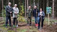 Eine Gruppe Menschen mit Spaten in den Händen stehen neben Jungbäumen, im Hintergrund Waldrand