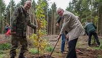 Zwei Männer, einer davon Soldat, beim Einsetzen eines Jungbaums, im Hindergrund weitere Menschen vor einem Waldrand