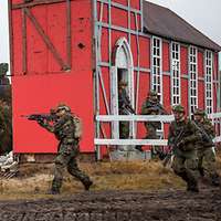 Fünf bewaffnete Soldaten laufen über ein Gefechtsfeld, im Hintergrund eine rote Gebäudeattrappe.