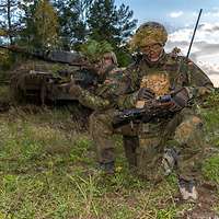 Zwei getarnte und bewaffnete Soldaten hocken an einem Waldrand, hinter ihnen steht ein Panzer.