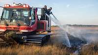 Ein Feuerlöschkraftfahrzeug fährt durch ein Moor, vom Heck aus löschen zwei Feuerwehrleute.