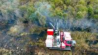 Drohnenaufnahme von einer Löschraupe, von der aus zwei Feuerwehrleute einen Brand im Moor löschen.