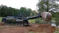 Ein Panzer mit einem Baggerarm schaufelt am Ufer Erde beiseite.
