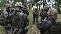 Eine Gruppe Soldaten steht in einem Laubwald im Kreis. Ein Soldat informiert alle anderen.