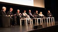 Acht Personen sitzen auf einem Podium und reden miteinander