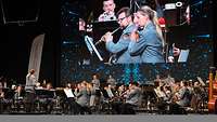 Musier spielen auf einer Bühne. Dahinter eine Leinwand mit Liveübertragung