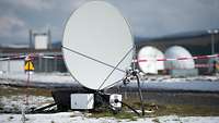 Vor einem rot-weißen Absperrband steht eine Satellitenschüssel in leicht schneebedecktem Gras.