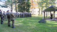 Soldaten stehen im Halbrund auf einer Rasenfläche unter Bäumen, vor ihnen steht ein Pfarrer.