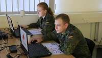 Eine Soldatin und ein Soldat sitzen in einem Büro vor zwei Laptops.
