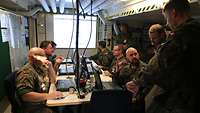 Mehrere Soldaten sitzen in einem Raum vor Computern und besprechen sich.