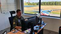 Bernd Göhler sitzt in seinem Büro am Flugplatz. Durch das Fenster sieht man ein Transportflugzeug.